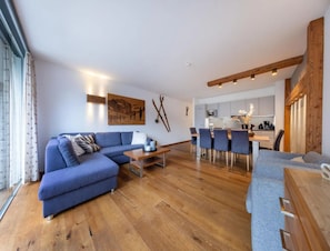 Un salon moderne avec un canapé d’angle, un coin repas avec une table et des chaises en bois, et une cuisine en arrière-plan.