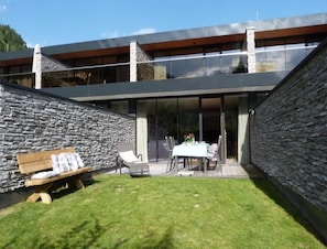 Une maison moderne avec une façade en pierre, un patio couvert avec une table et des chaises, et une cour gazonnée.