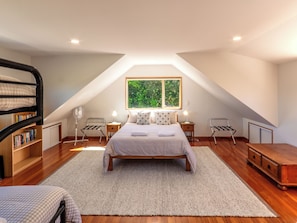 A bedroom with a sloped ceiling, a large window, a bed with a checkered bedspread, a bedside table with a lamp, a wooden dresser, and a bookshelf.