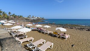 A beach with rows of sun loungers and umbrellas, a rocky shoreline, and a clear blue sky.