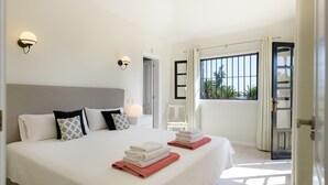 A hotel room with a large bed, two bedside tables, a nightstand, a window with curtains, and a view of palm trees.