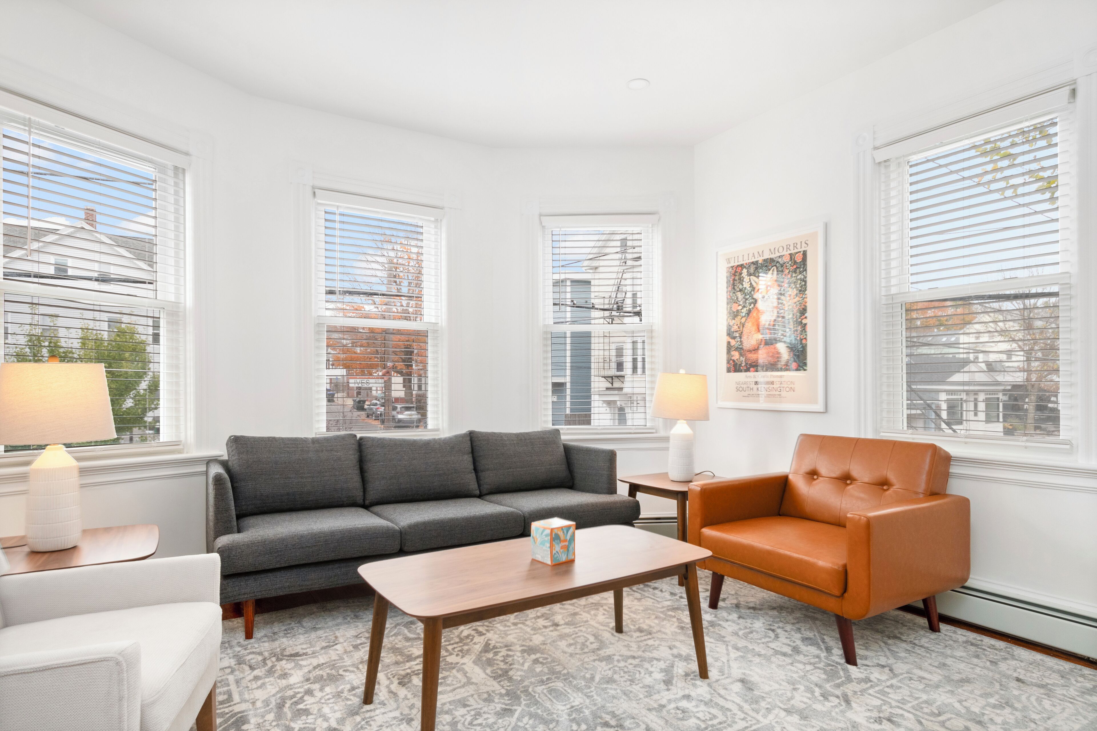 A modern living room with a gray sofa, an orange armchair, a wooden coffee table, and two side tables with lamps. Large windows with blinds provide views of the cityscape.