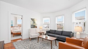 A spacious living room with a grey sofa, a brown leather armchair, a wooden coffee table, and a patterned rug. There is a white armchair, a small table with a lamp, and a painting on the wall. - Fox Point 3BR - Near Brown, RISD & Downtown (Providence)