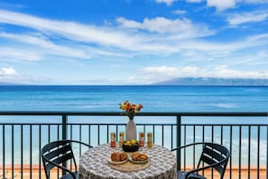 Una mesa redonda con mantel estampado, dos sillas, un florero con flores, un bol de croissants y dos latas de jugo, con vista al mar.