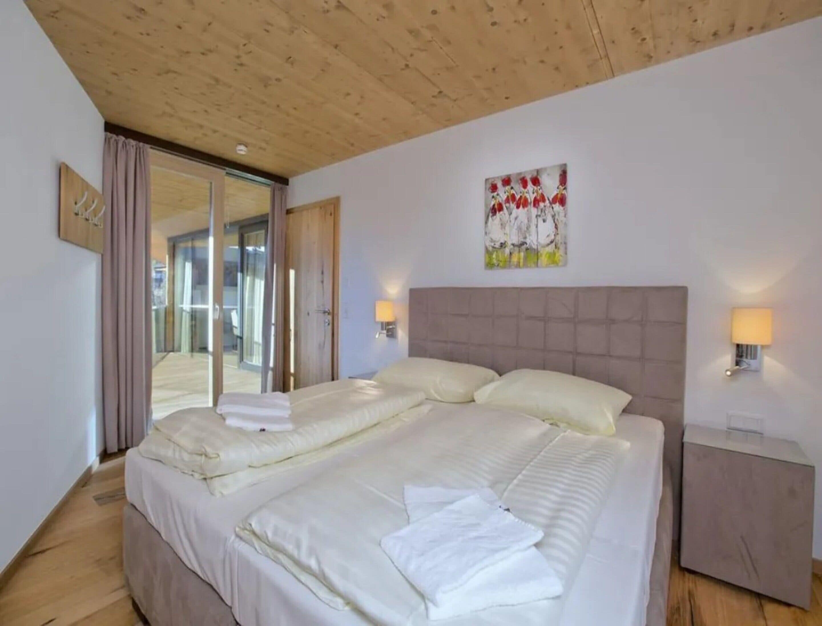 A hotel room with a double bed, a wooden ceiling, a glass door leading to a balcony, and a painting on the wall.