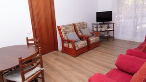 A living room with a wooden dining table, two floral-patterned chairs, a red sofa, and a television on a wooden stand. - APARTMENT GRANVIA FIRA BCN🎅🏻 (Hospitalet de Llobregat)