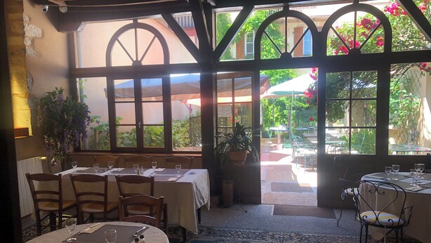 A restaurant with a rustic interior, featuring wooden beams and large windows with a view of the outdoor garden. Tables are set with white tablecloths, and there are potted plants and a patterned rug on the floor. - Hôtel de l'Europe (Corbigny)
