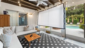 A modern living room with a white sofa, wooden coffee table, and a ceiling fan. A large window offers a view of a pool and greenery.