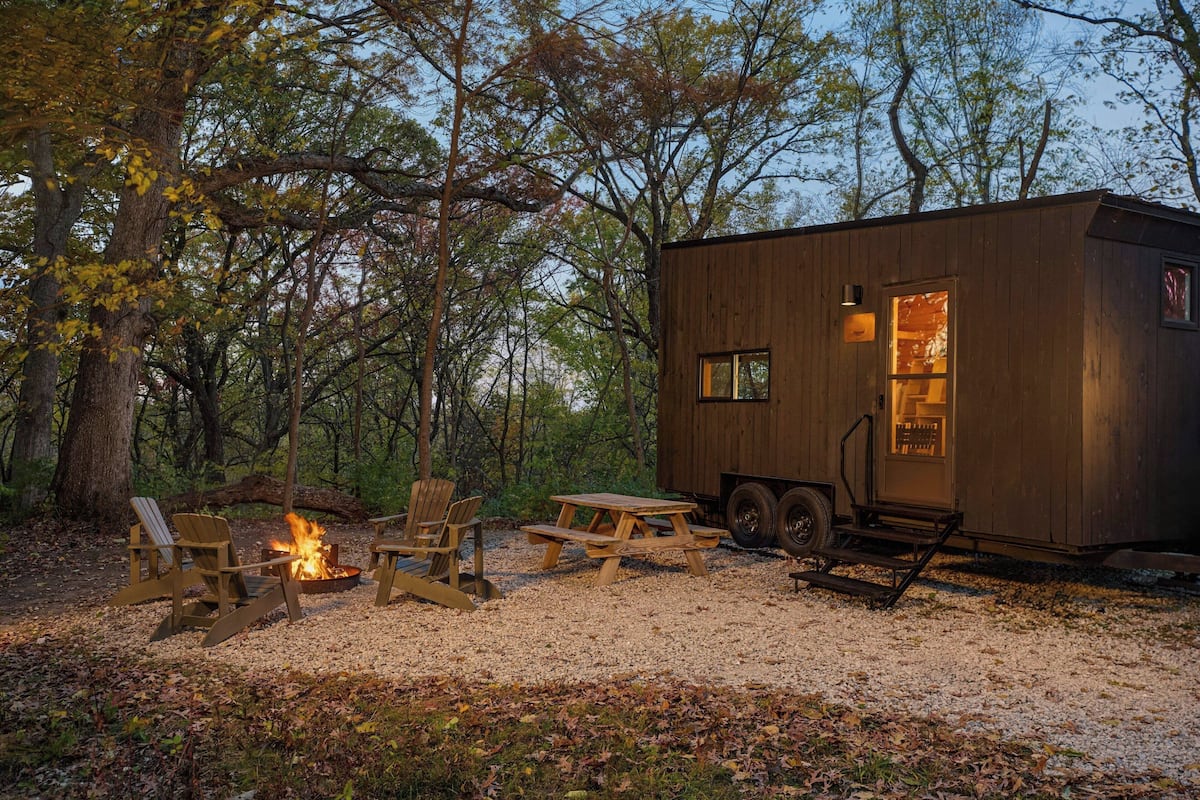 A small wooden cabin with a fire pit, picnic table, and two chairs in a forest setting.