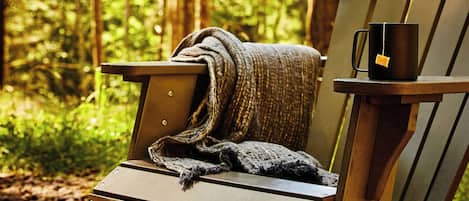 A wooden Adirondack chair with a blanket and a mug in a forest setting.