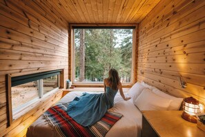 A wooden cabin bedroom with a large window, a bed with a patterned blanket, and a nightstand with a lantern. - Postcard Cabins Big Bear, Outdoor Collection by Marriott Bonvoy (Running Springs)