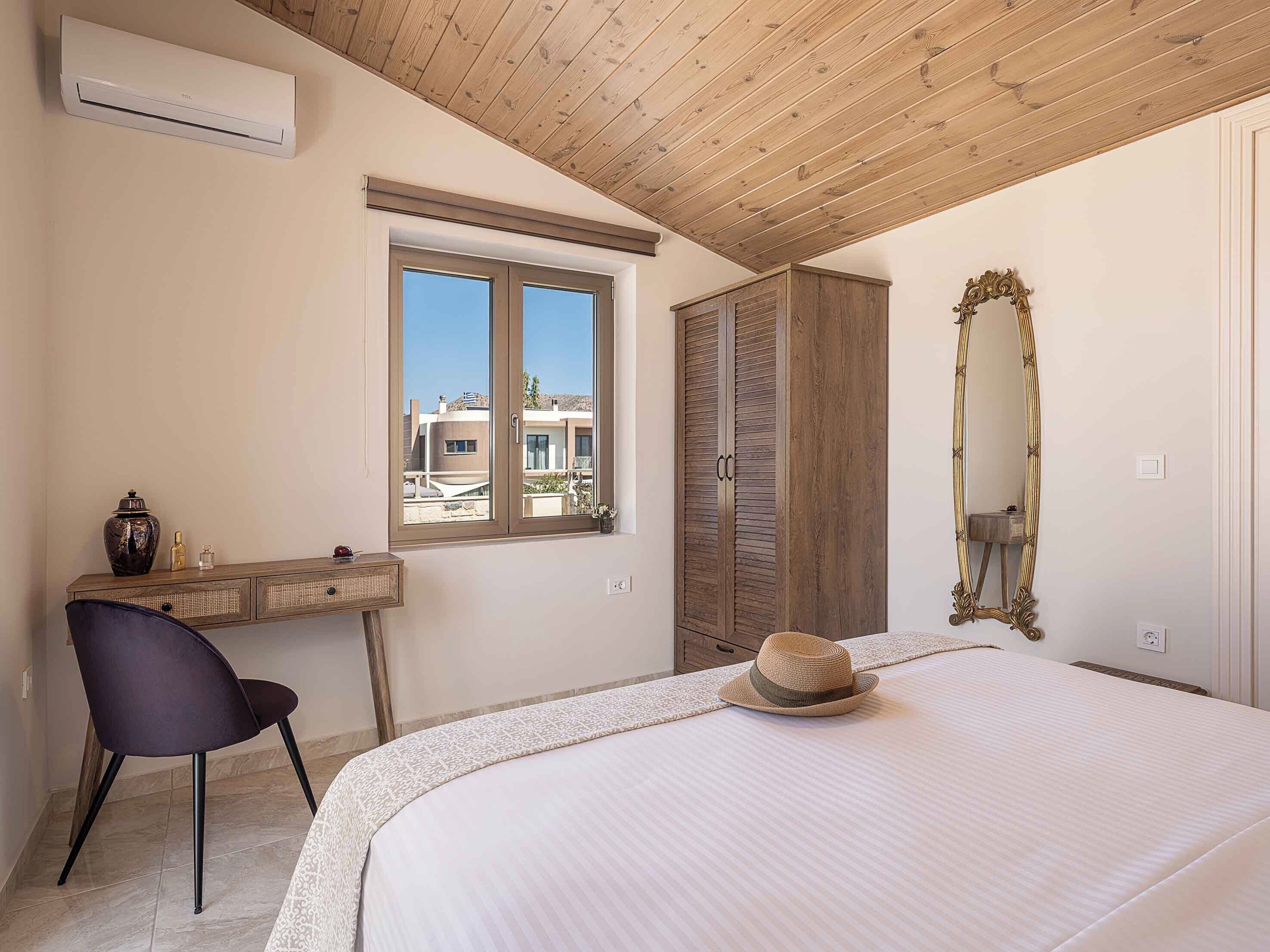 Ein Schlafzimmer mit Bett, Schreibtisch, Stuhl, Kleiderschrank, Spiegel und einem Fenster mit Blick auf Gebäude.