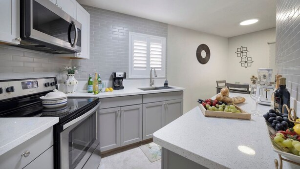 Una cocina moderna con armarios blancos, una estufa de acero inoxidable y un microondas. Hay un frutero en la encimera y una cafetera.