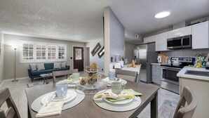 Una cocina moderna con electrodomésticos de acero inoxidable, una zona de comedor con una mesa redonda y una sala de estar con sofá y ventana con persianas.
