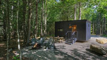 Una cabaña en un bosque con un camino de grava, terraza de madera y sillas Adirondack.