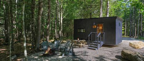 Een chalet in een bos, met een grindpad, een houten terras en Adirondack-stoelen.