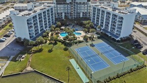 Un hotel de varios pisos con un patio central, piscina al aire libre, canchas de tenis y vista al mar.