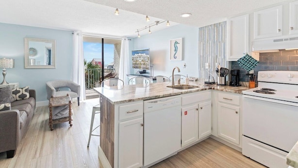 Una cocina moderna con armarios blancos, una isla central con un fregadero y una estufa. Hay una zona de comedor con un sofá gris y una pequeña mesa de madera.