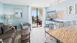 Una sala de estar con sofá, sillón y mesa de comedor. Un puerta corredera da acceso a un espacio exterior con vistas a edificios.