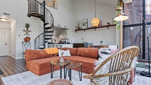 A spacious living area with a large sectional sofa, a round wooden coffee table, and a wicker chair. A spiral staircase leads to an upper level with a kitchen and dining area.