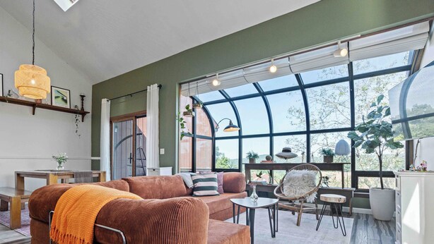 A spacious living room with a large window, a brown sofa, a dining table, and a hanging light fixture.