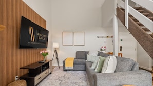Un salon moderne avec un canapé gris, un meuble de rangement en bois, un téléviseur à écran plat, un escalier et un tapis à motifs.