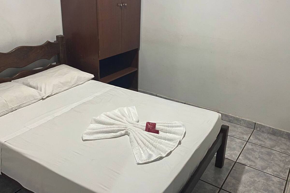 A neatly made bed with a towel arranged in a fan shape, a wooden wardrobe, and a tiled floor.