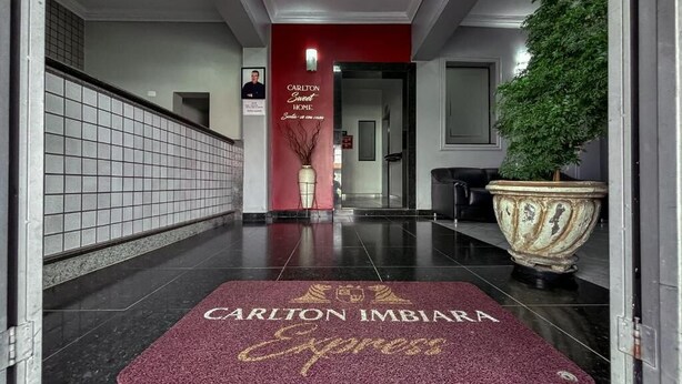 L’entrée d’un hôtel, avec un mur rouge, une grande plante en pot et un paillasson sur lequel on peut lire : « Carlton Imbira Express ».
