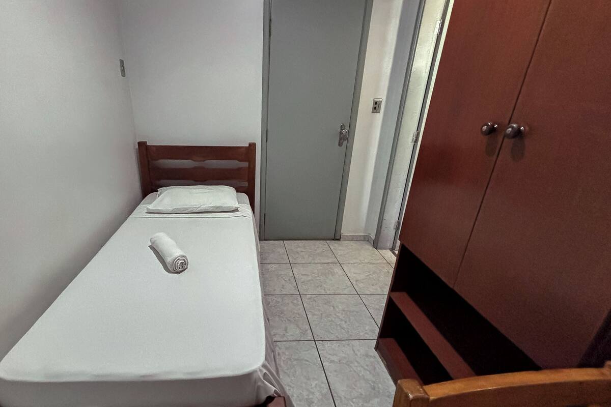 A small, narrow room with a single bed, a wooden wardrobe, and a tiled floor.