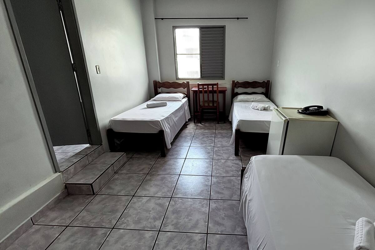 A small, narrow room with two single beds, a desk with a chair, and a window with blinds.