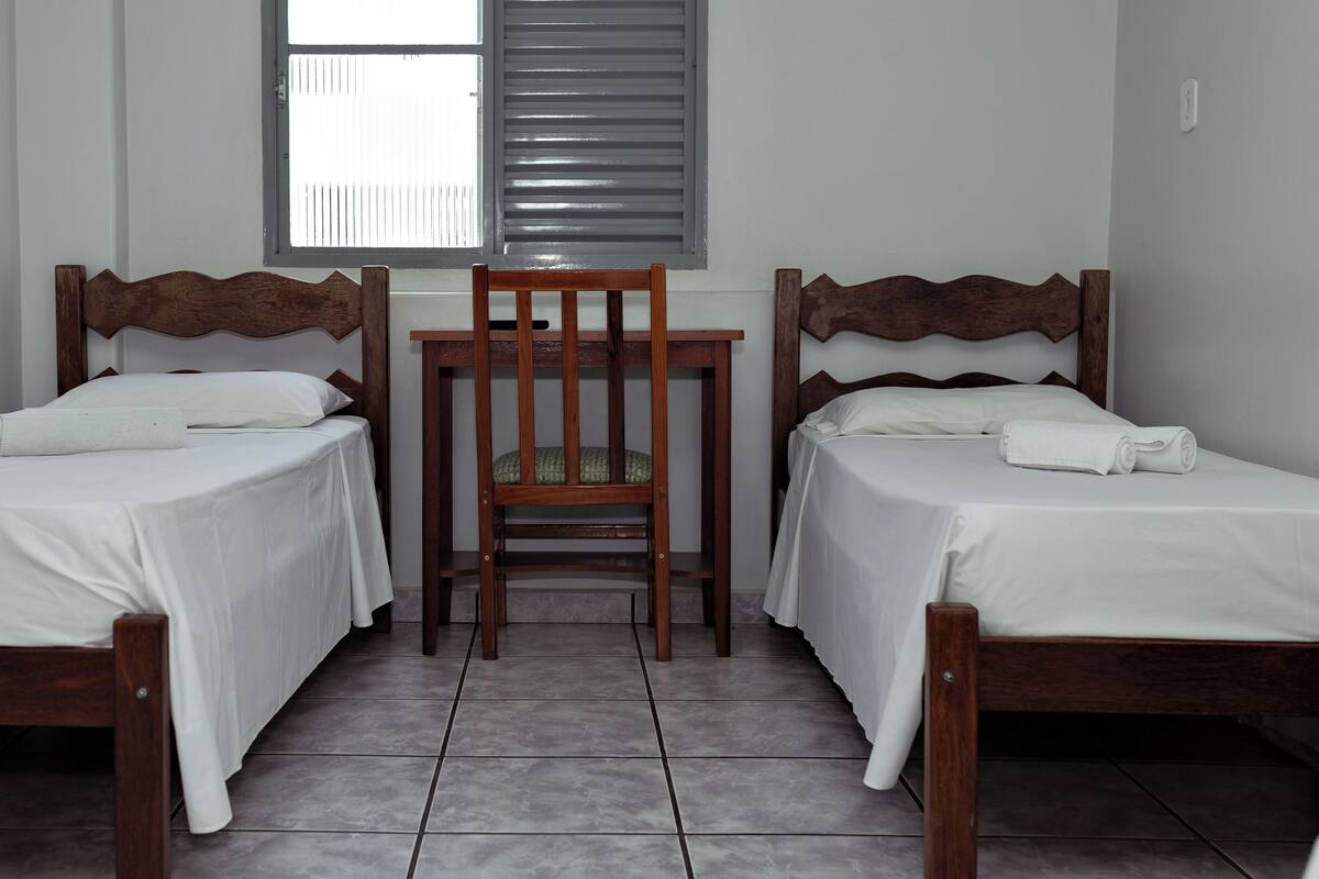 A room with two wooden beds, a small table, and a window with blinds.