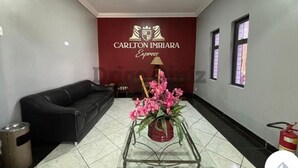 A hotel lobby with a black leather sofa, a glass coffee table with pink flowers, and a red wall with the Carlton Imbira Express logo. - Hotel Carlton Imbiara Express (Araxá)