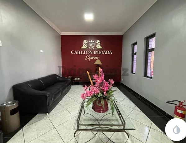 A hotel lobby with a black leather sofa, a glass coffee table with pink flowers, and a red wall with the Carlton Imbira Express logo. - Hotel Carlton Imbiara Express (Araxá)