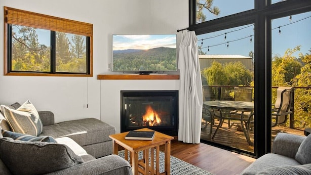 A modern living room with a fireplace, comfortable seating, a television, and a balcony with a scenic view. - YOLO Lake Escape | Lakefront Views - EV Charger (Big Bear Lake)