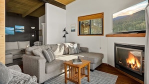 A modern living room with a grey sofa, a fireplace, a television, and a large window with a forest view. - YOLO Lake Escape | Lakefront Views - EV Charger (Big Bear Lake)
