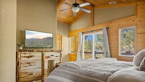 A bedroom with a bed, wooden dresser, television, and large windows with curtains. - Enchanting Escape | Hot Tub, Fire Pit & Game Room (Big Bear Lake)