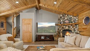 A cozy cabin interior with a stone fireplace, a television mounted on the wall, and a comfortable seating area with sofas and a coffee table. - Enchanting Escape | Hot Tub, Fire Pit & Game Room (Big Bear Lake)