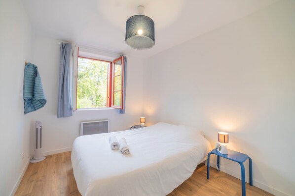 Une chambre à coucher avec un lit, une fenêtre avec des rideaux, un radiateur mobile, une table de chevet avec une lampe et un luminaire au plafond.