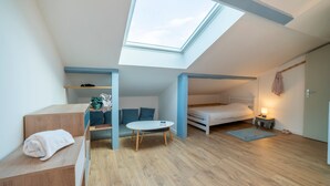 Habitación compacta y moderna con tragaluz, una pequeña mesa redonda, un sofá, una cama y una mesita de noche con lámpara.