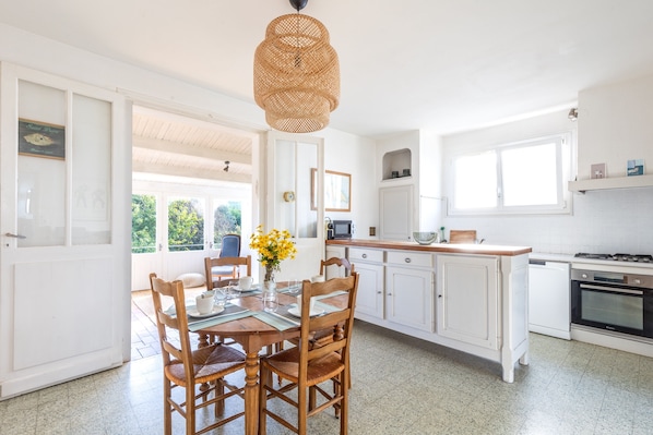 A bright kitchen with white cabinetry, a dining table with chairs, a hanging light fixture, and a view through the open door to the outside.
