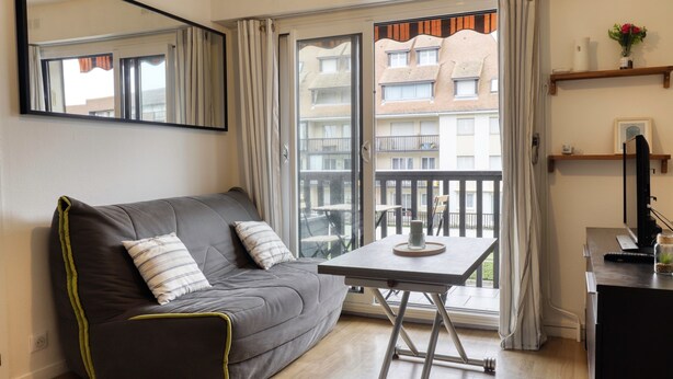 A modern living room with a grey sofa, a small table, a TV, and a balcony with a view of buildings. - L'Estran - Appt 2p, proche de la mer (Villers-sur-Mer)