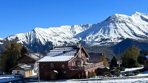 A wooden chalet with a balcony, set against a backdrop of snow-covered mountains.