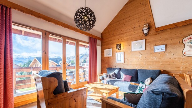 A cozy wooden interior with a sofa, chairs, and a table. Large windows offer a view of the outdoors.