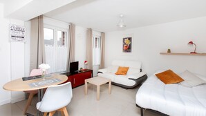 Una sala de estar moderna con un sofá blanco, una pequeña mesa de madera, un televisor sobre un mueble rojo y un comedor con una mesa y sillas.