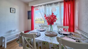 Un coin repas avec une table dressée pour deux personnes, un vase rempli de fleurs rouges et une vue sur l’extérieur grâce à de grandes fenêtres ornées de rideaux rouges.