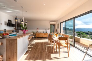 A modern kitchen with a wooden island, dining area with wooden chairs, and large windows offering a view of the mountains. - Maison Altea - Maison climatisée (Figari)