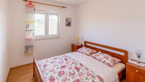 A bedroom with a wooden bed, floral bedding, a bedside table with a lamp, a window with blinds, and a wall-mounted shelf with colorful containers. - One Bedroom Apartment with Balcony (Okrug Gornji)