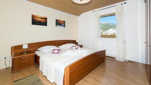 A bedroom with a wooden bed, a nightstand, a lamp, a window with curtains, and two wall-mounted pictures. - Four Bedroom Holiday Home with Pool (Kastel Stari)