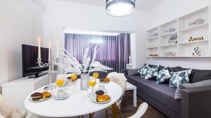 Un salon moderne avec une table à manger dressée pour le petit-déjeuner, un canapé agrémenté de coussins décoratifs et un téléviseur posé sur un meuble.