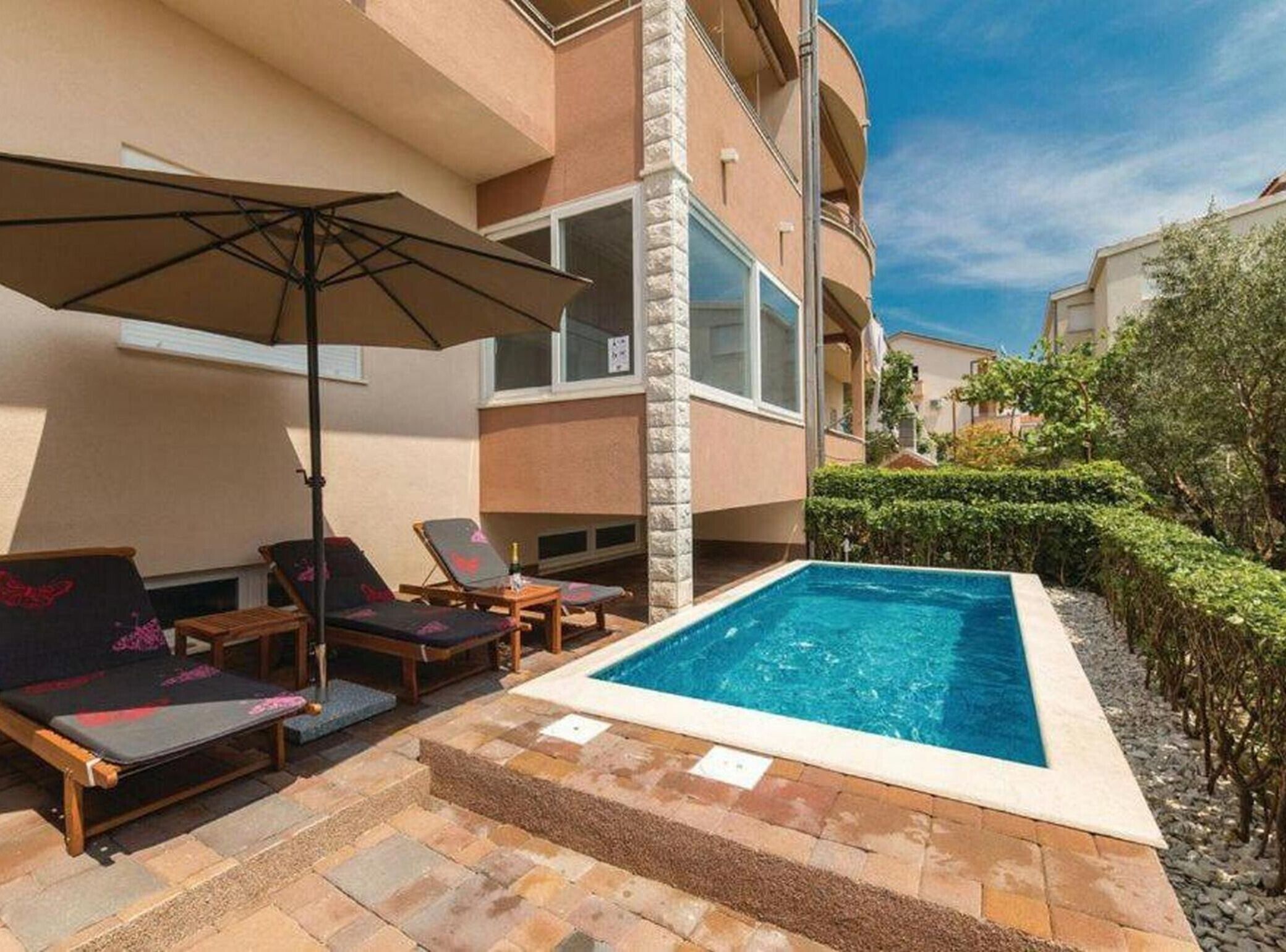 Un espace piscine doté de chaises longues et d’un parasol, entouré d’une terrasse en pierre et d’un bâtiment avec des balcons.